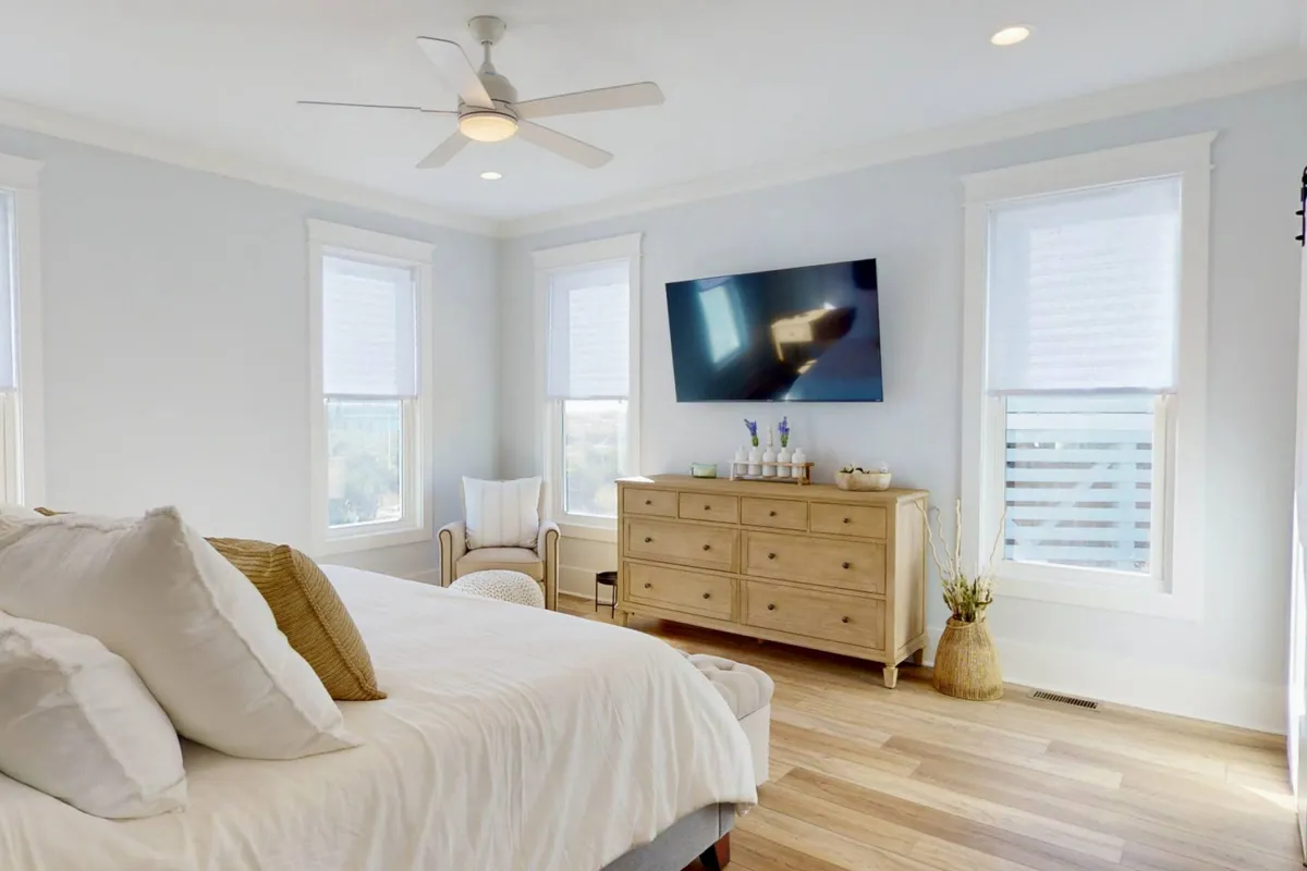 An inviting king bedroom featuring plush pillows, a wooden dresser, a TV on the wall, and a comfortable chair. Sunlight streams into the room, creating a cozy and welcoming environment.