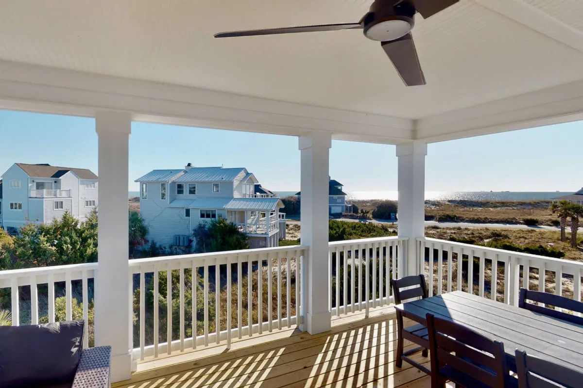 A serene outdoor dining space with a view of the ocean. The table is set for al fresco meals, and the covered patio includes a ceiling fan, with the sea and surrounding homes in the background. The space is clearly designed for gatherings, relaxation, and enjoying the coastal atmosphere.