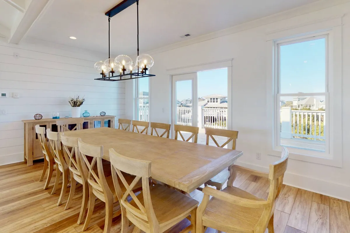 A large wooden dining table under a beautiful chandelier, surrounded by comfortable seating and set near windows that offer glimpses of the outdoors. The setup invites group meals and celebrations, with an inviting and communal mood.