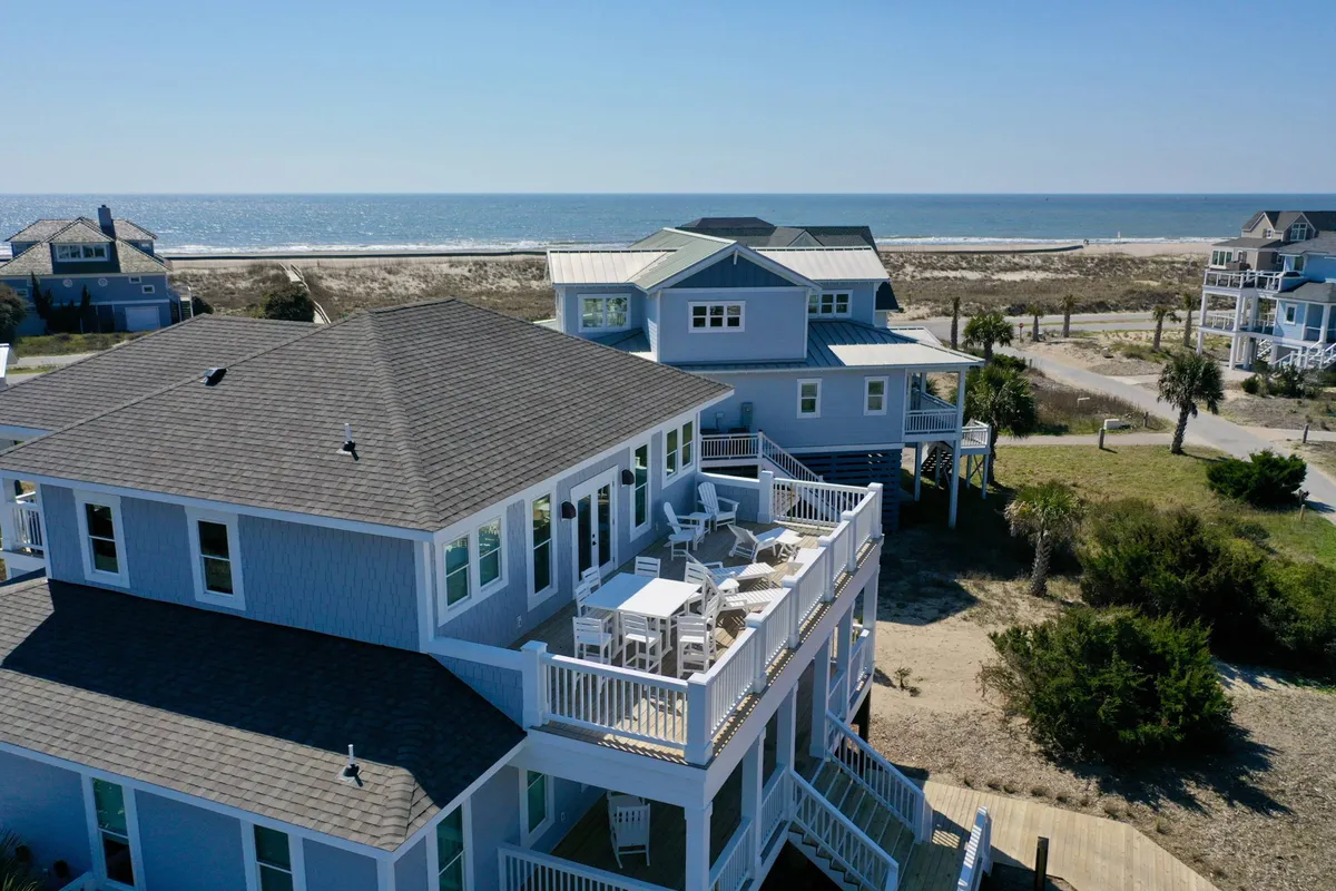 An expansive beachfront deck with comfortable outdoor seating overlooks the ocean, surrounded by lush greenery and framed by railings. The space is designed for gatherings, outdoor dining, and watching sunsets together as a group.