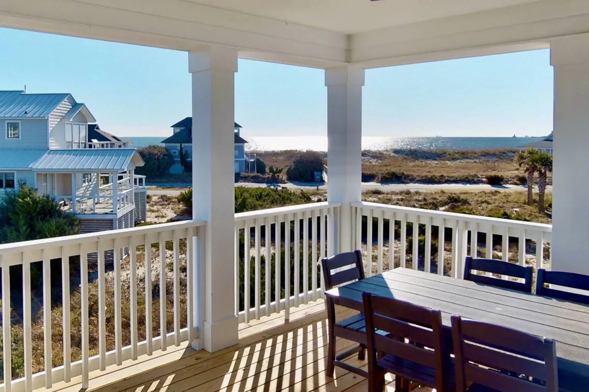 The image showcases an inviting outdoor dining area overlooking sweeping ocean views, with the house's elegant exterior and a backdrop of neighboring homes. The space is set up for gatherings, with a large table perfect for sharing meals, relaxing, and enjoying the stunning sunsets over Bald Head Island.