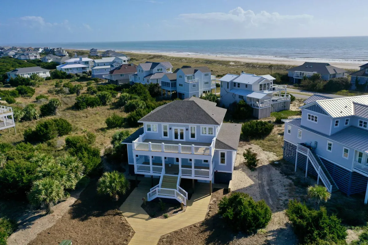 A wide beachfront view of the property’s main entrance and deck, with the beach and ocean stretching beyond. The deck is equipped for group gatherings and outdoor dining, emphasizing the home’s location right on the sand.