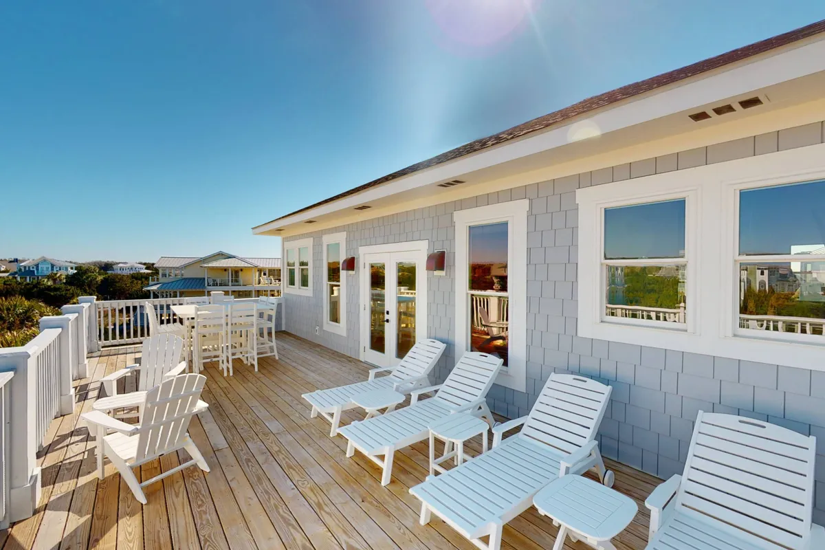 A spacious outdoor deck featuring lounge chairs, a dining area, and large windows that hint at the seamless flow between interior and exterior spaces. The deck offers direct ocean views and is set up for relaxation, socializing, and outdoor meals.