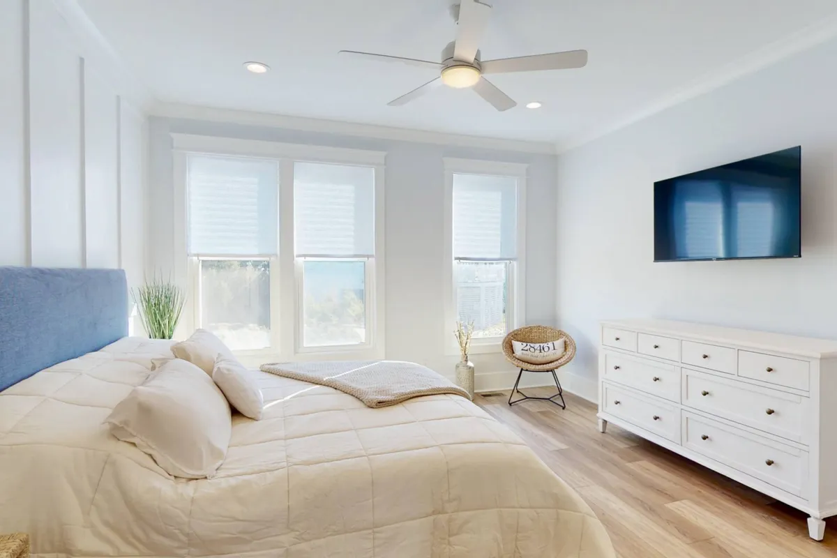 A serene and spacious king bedroom with a large bed, bright windows letting in natural light, a television, and a dresser. The mood is tranquil and the setting promises comfort and relaxation.