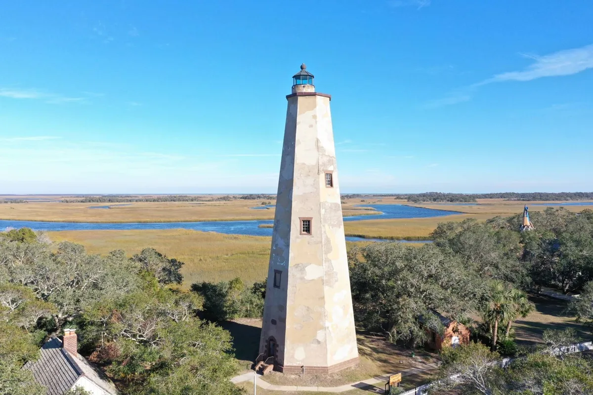 The iconic Old Baldy Lighthouse stands tall against a blue sky, surrounded by marshland. The foreground features natural grasses and walking paths, inviting exploration and discovery—ideal for family outings and local adventure.