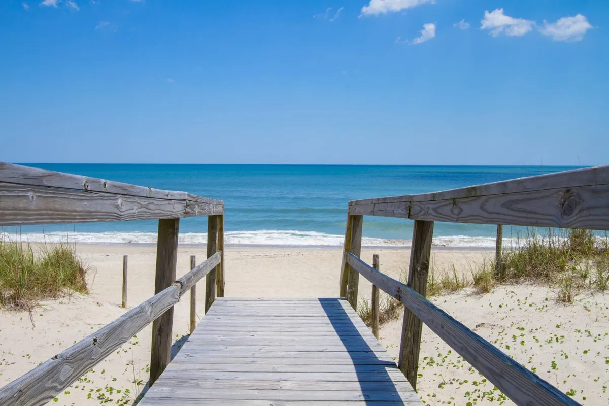 A wide wooden walkway leads directly to the sandy beach, with panoramic views of the ocean and a clear blue sky stretching overhead. The scene captures a tranquil, open shoreline perfect for sandcastle-building, morning walks, and family fun by the water.