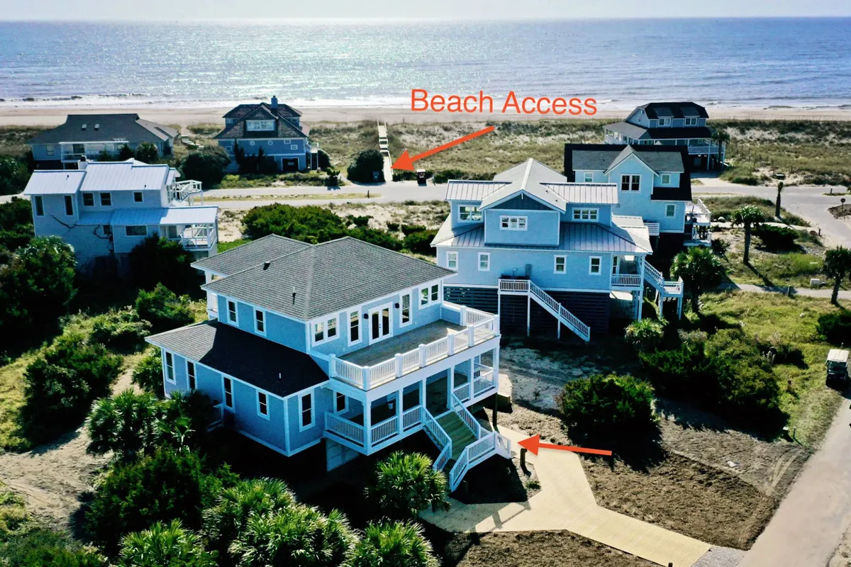 A welcoming exterior scene featuring the main house set amongst lush greenery, with a sandy pathway leading directly to the beach. The foreground highlights easy beach access, while the home’s decks suggest opportunities for sunset dinners and relaxation overlooking the ocean.