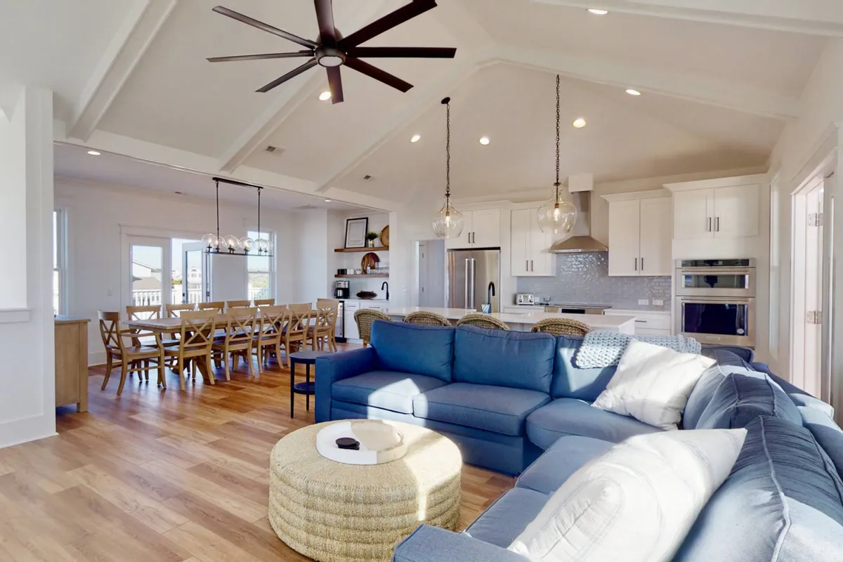 A bright, open-concept interior showing a spacious sectional couch arranged around a round coffee table, with a dining table and chairs positioned nearby. The open kitchen is visible in the background, seamlessly connecting the living, dining, and kitchen spaces. Large windows allow natural light to pour in, and the overall setting feels airy and inviting, perfect for gatherings.